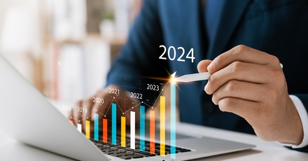 The image depicts a businessperson using a laptop, with a pen pointing towards a bar chart projected in front of the screen. The chart shows a rising trend in data from 2019 to 2024, with the years marked on the bars, indicating growth or progress over these years. The person is in formal attire, suggesting a professional or business context.